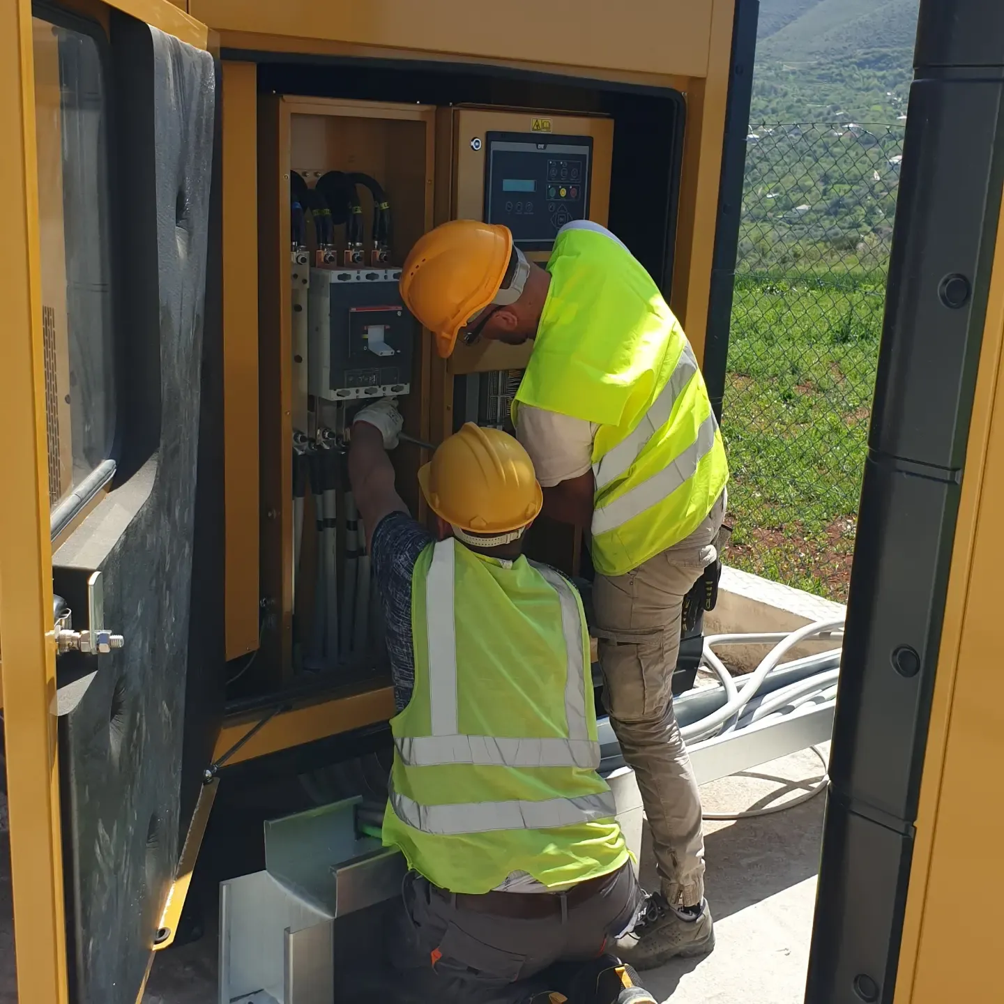Dy persona me helmeta dhe jelekë reflektues punojnë në panelin elektrik të një kabine të jashtme të verdhë.
